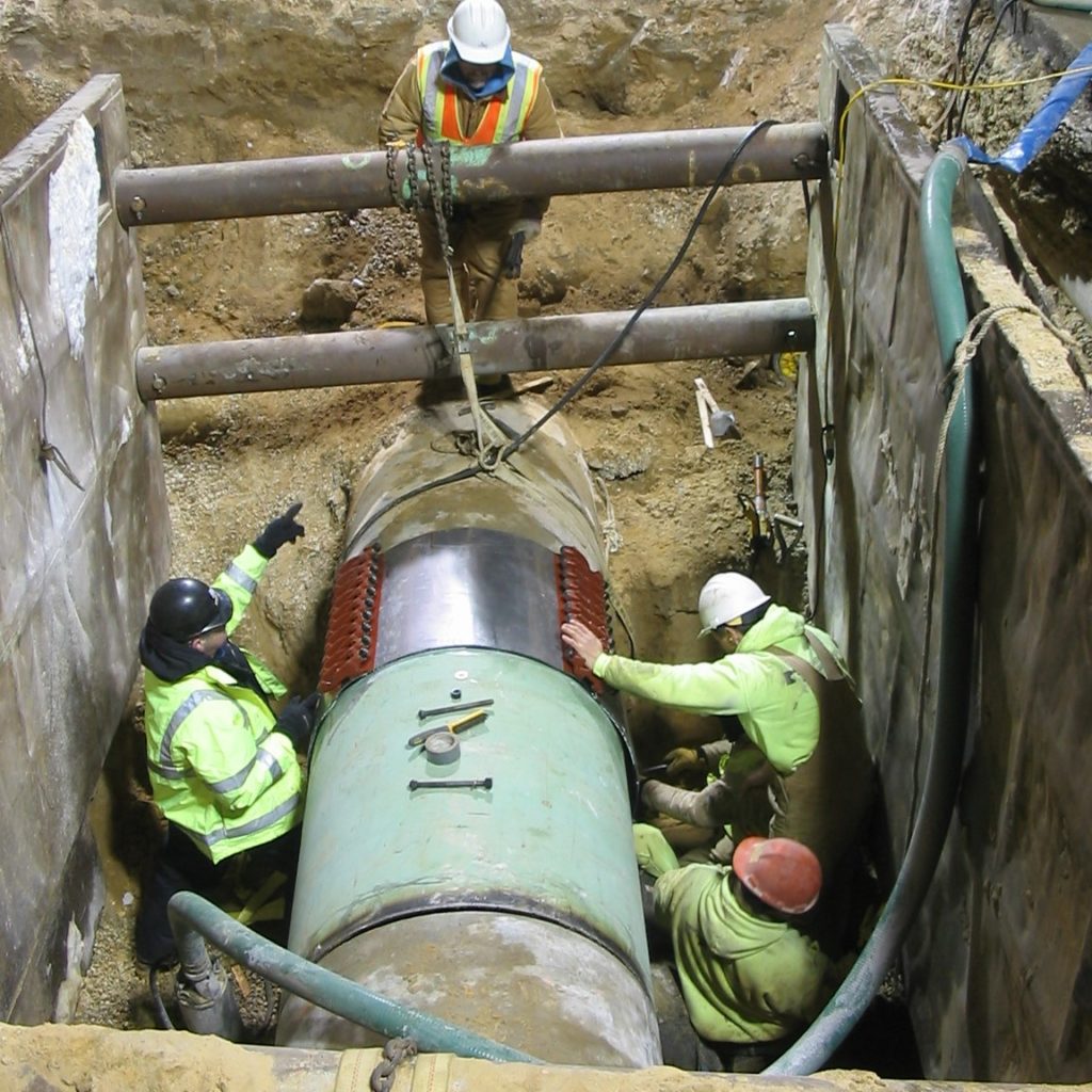 Workers in hard hats install a replacement section of forcemain pipe and add clamps to seal the joints.