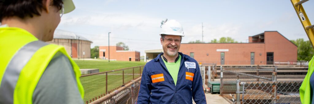 A plant operator smiles with wastewater treatment tanks and buildings in the background while talking to an engineer.