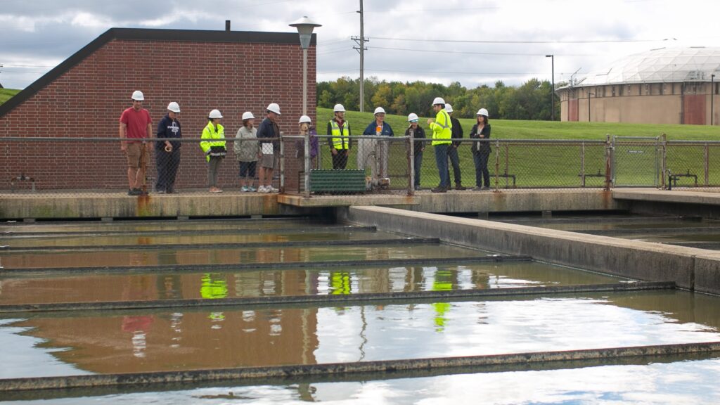 A tour group in safety helmets stand in front of wastewater tanks to learn about processes and operations of sewage treatment.