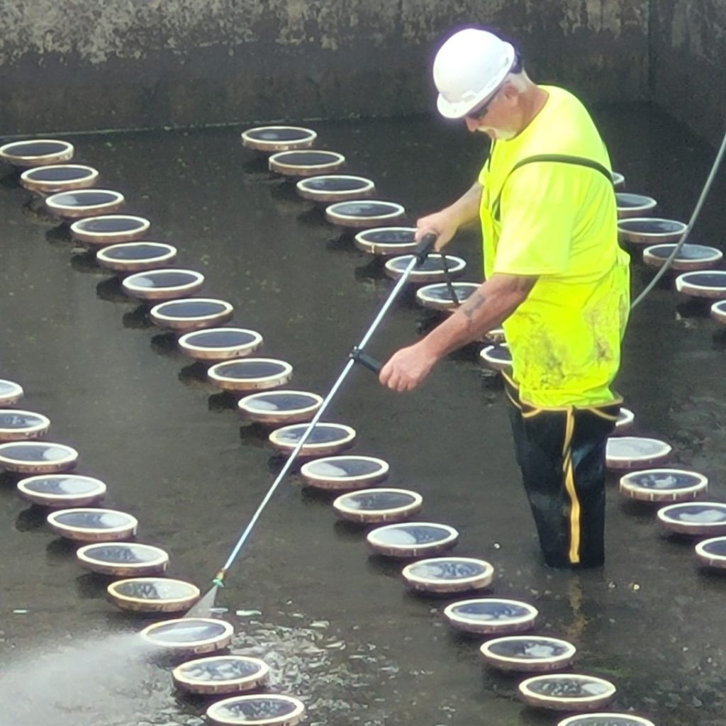 A staff member in high vis waders power washes aeration tank diffuser stones.