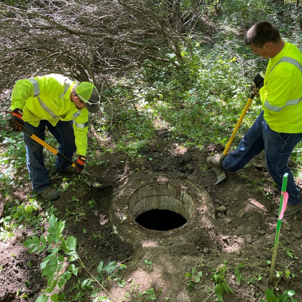 Two men dig out a manhole frame to expose concrete rings.