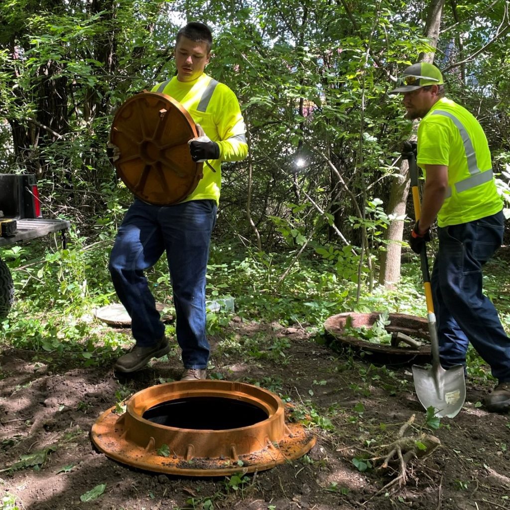 One man places the manhole cover back on top of the casting while another man works to bury the casting.