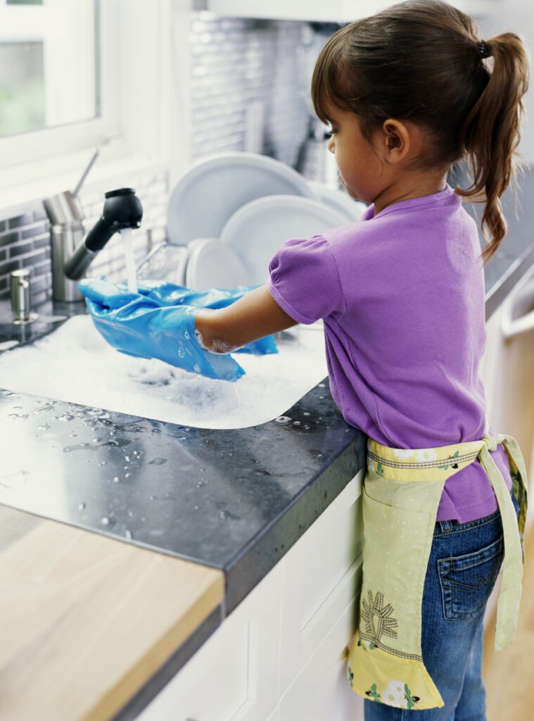 A little girl washes dishes wearing gloves and an apron at a kitchen sink.