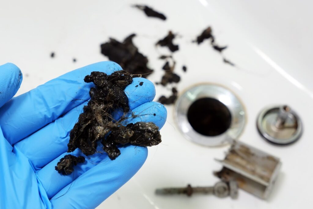 A gloved hand hold a mixture of hair and grime that was pulled out of a clogged drain. In the background shower or sink drain plumbing parts are visible.