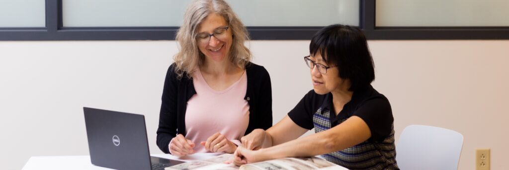 Two female office staff work at a computer and look at a photo album.