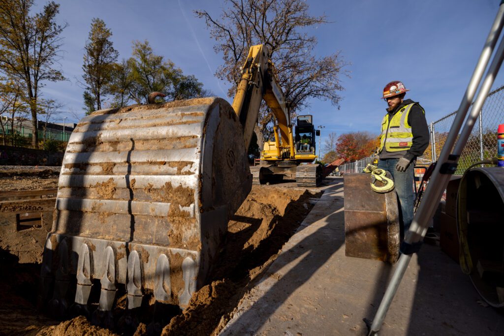 A photo of an excavator digging at a construction site with a worker overlooking the equipment and work.
