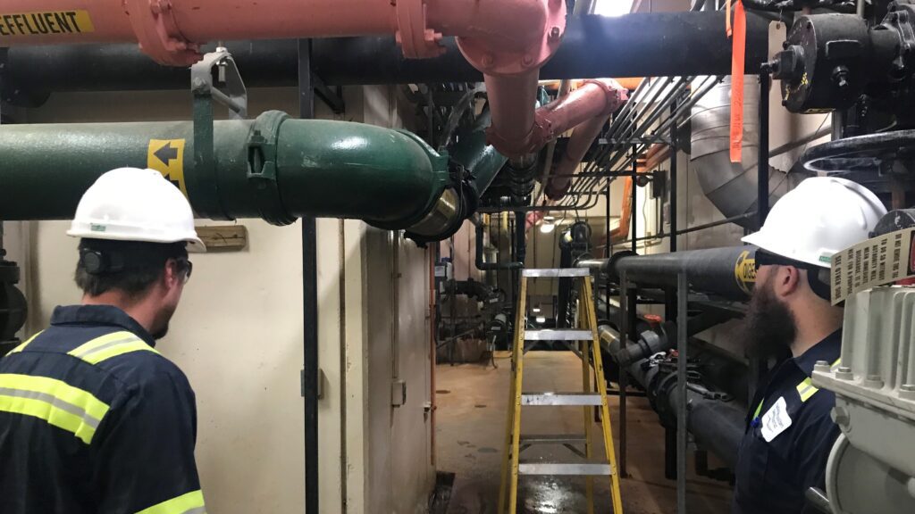 Two mechanics in hard hats look at pipes and valves at the treatment plant.