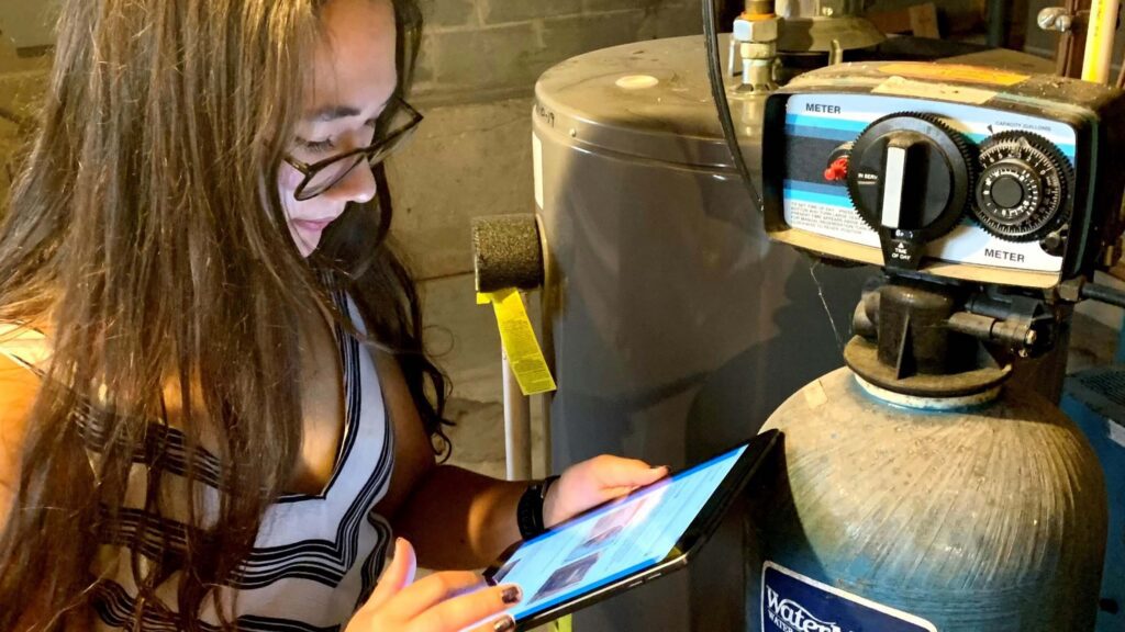 A young woman looks at a tablet in front of her aging water water softener.