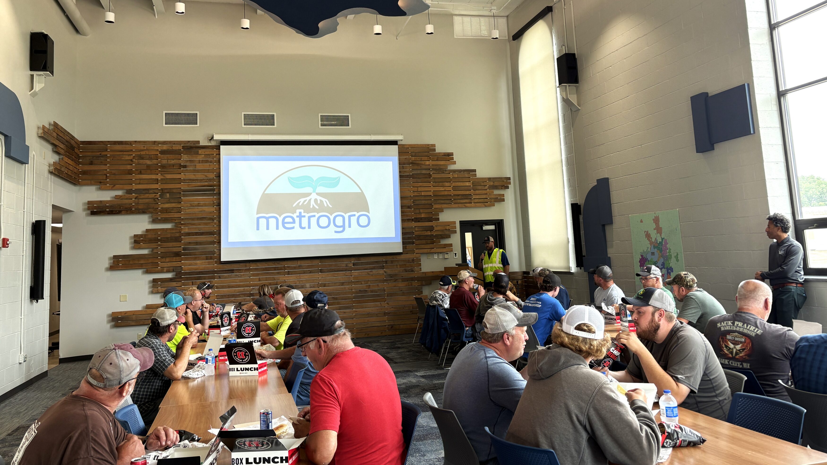 A District staff member stands facing a room of attendees eating lunch at tables. A large screen behind them displays the Metrogro logo.